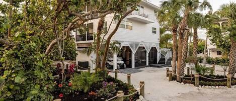 A white building with a covered outdoor area, surrounded by palm trees and a garden with various plants and flowers.