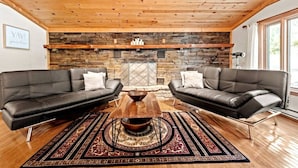 A cozy living room with two leather sofas, a wooden coffee table, and a patterned rug. A stone fireplace with a metal screen is in the background.