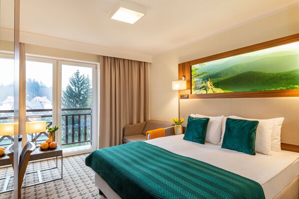 A hotel room with a large bed, a view of a balcony, a painting of a landscape, and a table with a lamp and oranges.
