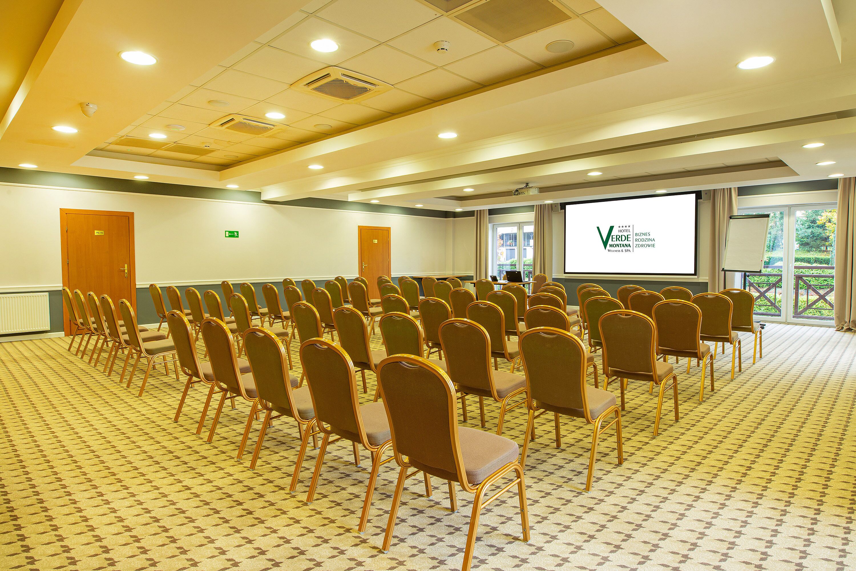 A conference room with rows of chairs, a large screen, and a view of greenery outside.