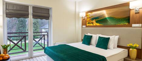 Habitación de hotel con cama, un cuadro en la pared, un balcón con vistas a vegetación y una mesa con fruta.