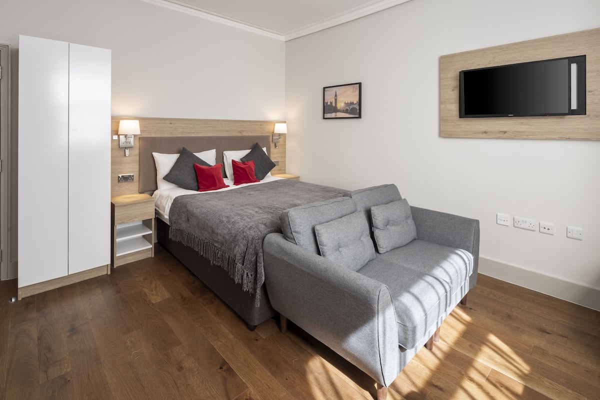 A modern hotel room with a bed, grey sofa, wooden floor, and a flat-screen TV mounted on the wall.