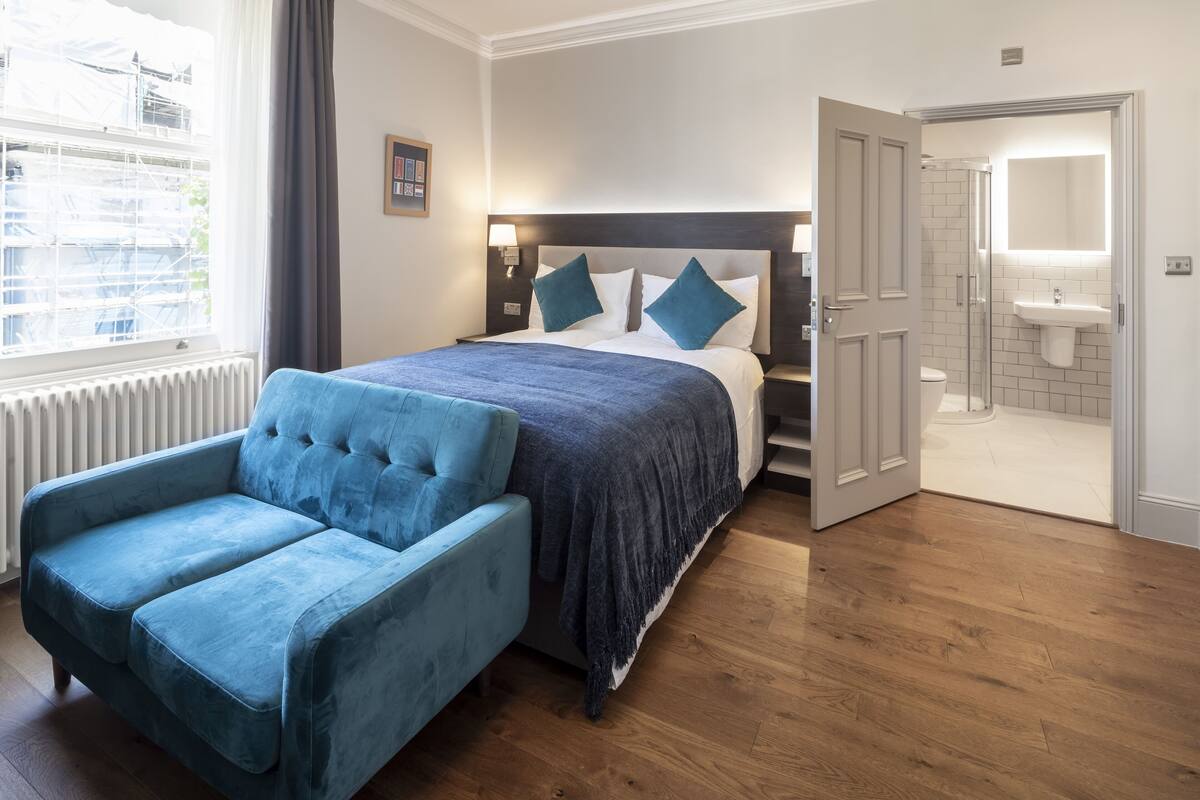 A bedroom with a bed, a blue armchair, a nightstand, a bedside table, a window with curtains, a door to a bathroom, and a view of the outside.
