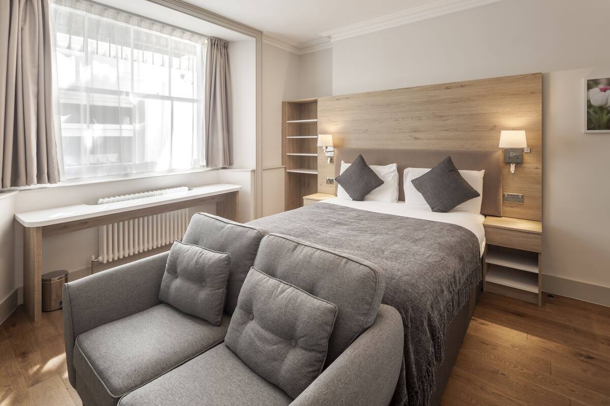 A modern hotel room with a large bed, grey upholstered chairs, a wooden headboard, bedside lamps, and a radiator.