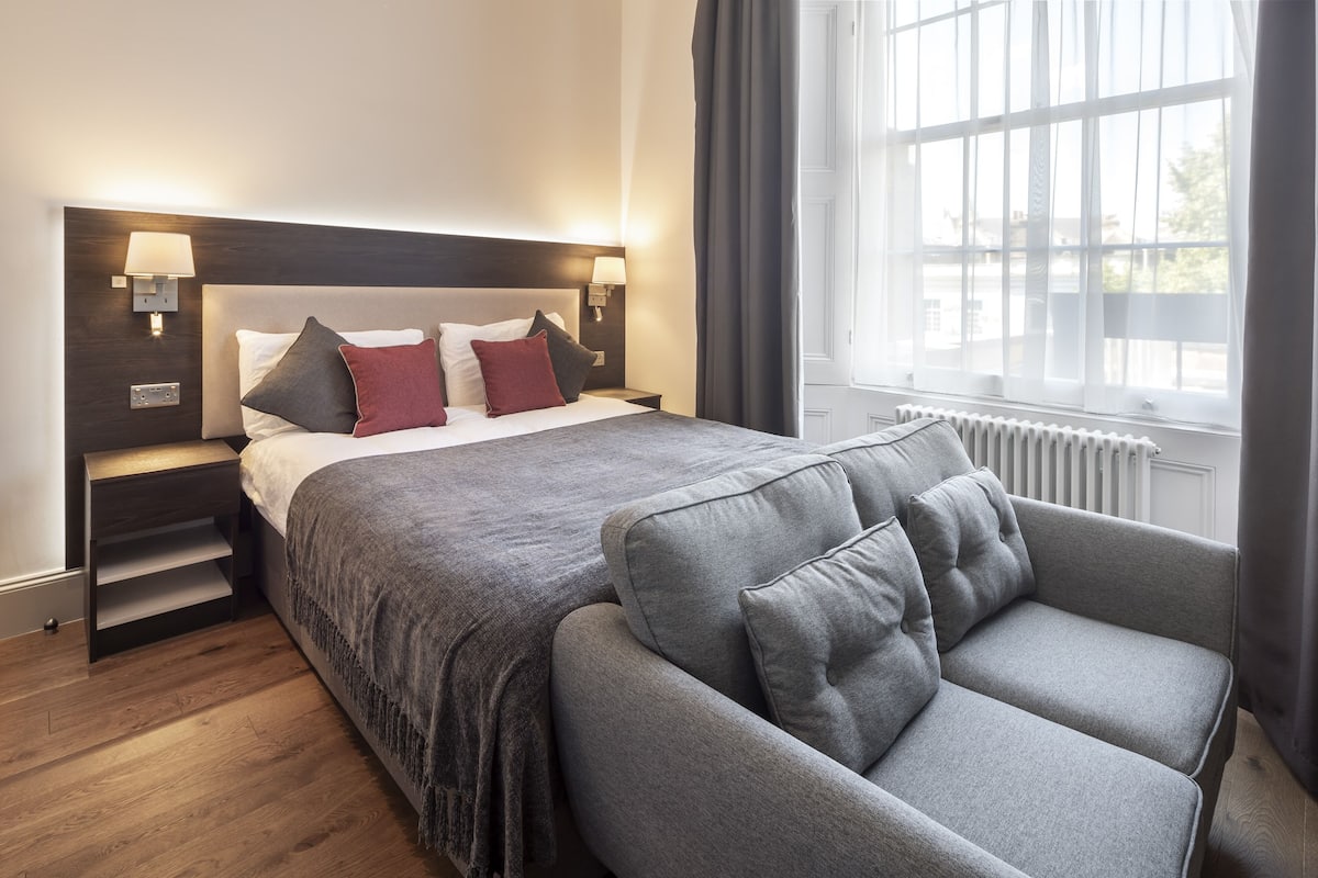 A modern hotel room with a large bed, grey upholstered armchair, and a view of the outdoors through a window.