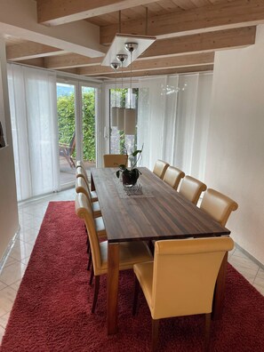 A dining area with a wooden table and beige chairs, a red rug, and a view of greenery through large windows.