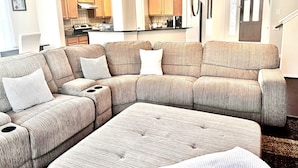 A living room with a sectional sofa, ottoman, and a kitchen area in the background.