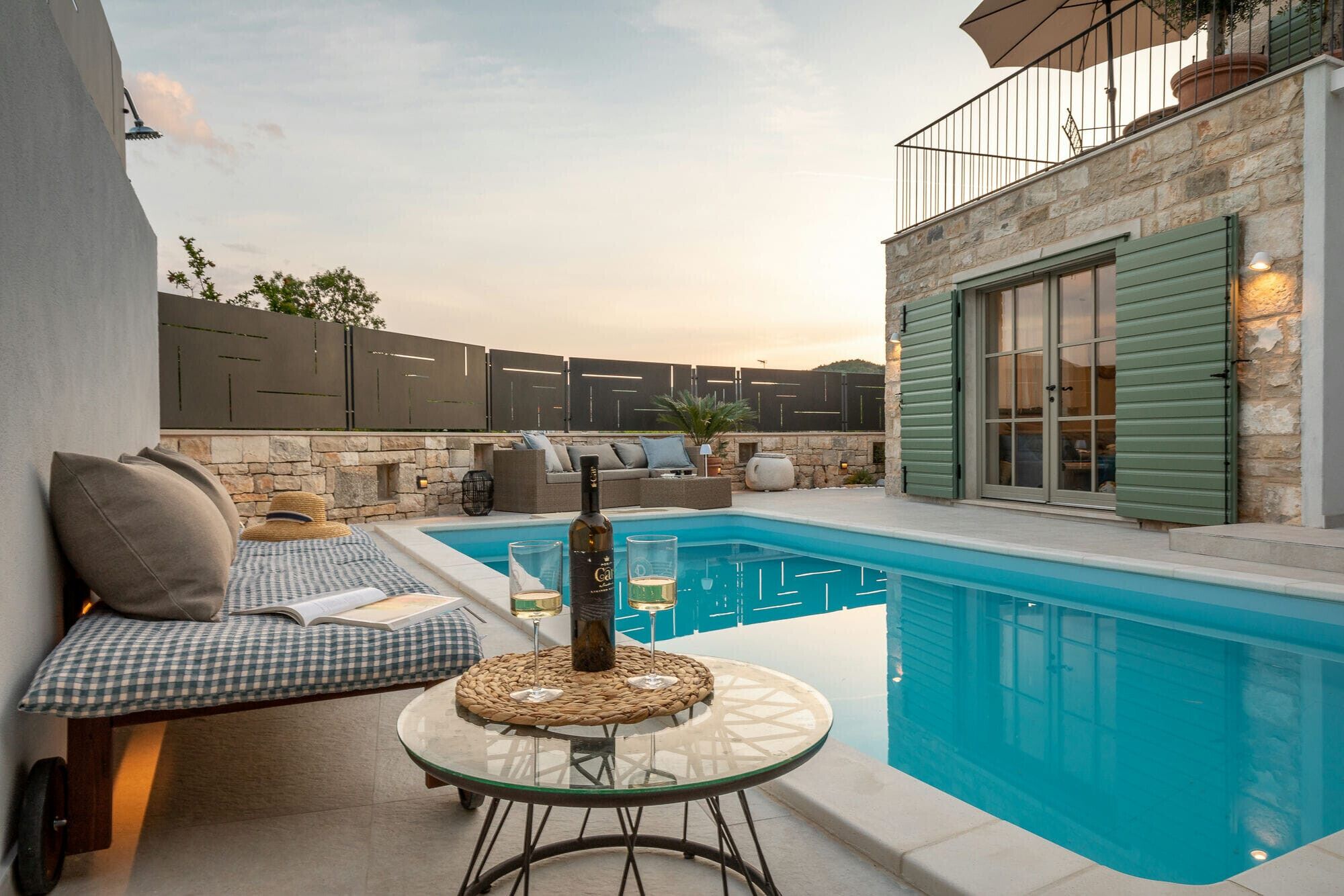 A pool area with a glass table, a bottle of wine, and a sofa with cushions.