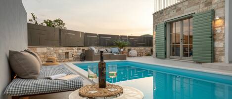A pool area with a glass table, a bottle of wine, and a sofa with cushions.