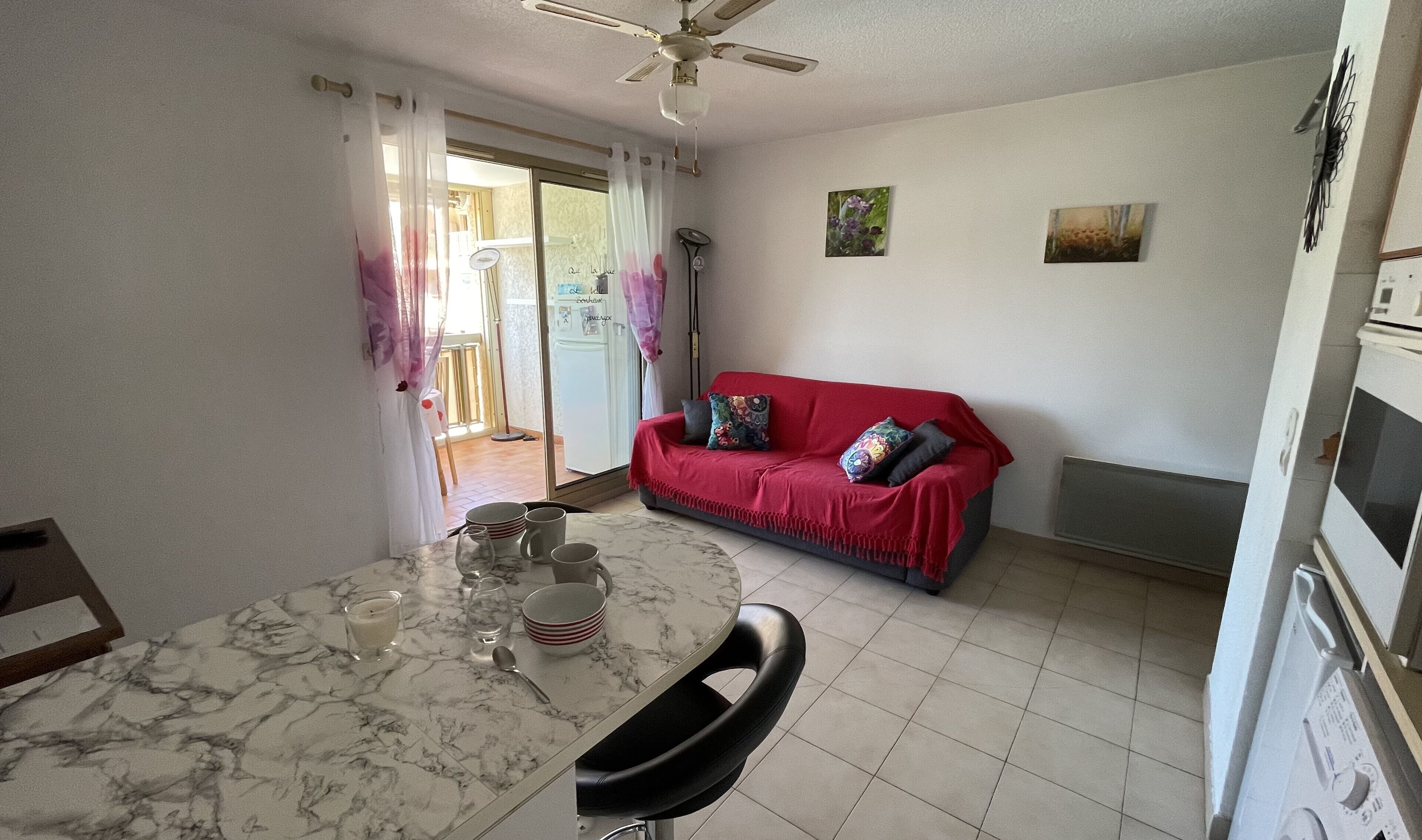 A compact living space with a dining area featuring a marble table and chairs, a red sofa with cushions, and a ceiling fan.