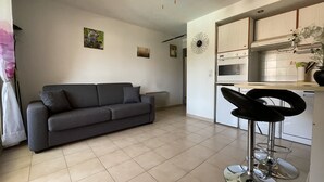 A modern living room with a grey sofa, a kitchen area with a white oven and microwave, and a bar stool. - T2 with balcony close to the sea. (Sainte-Maxime)