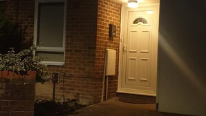 Vue nocturne de l’entrée d’un immeuble, avec une porte éclairée et une petite fenêtre sur la gauche.