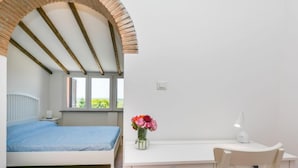 Ein Schlafzimmer mit Bett, Schreibtisch und Stuhl. Es gibt ein Fenster mit Jalousien, eine Blumenvase und eine Lampe auf dem Schreibtisch.