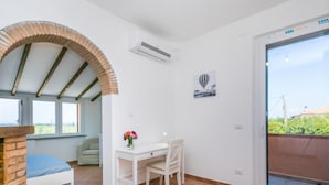 Ein Zimmer mit weißem Schreibtisch, einem Sessel, einem kleinen Tisch mit Blumenvase, einem Kamin aus Ziegelsteinen, einem großen Fenster und einer Schiebetür zu einem Außenbereich.