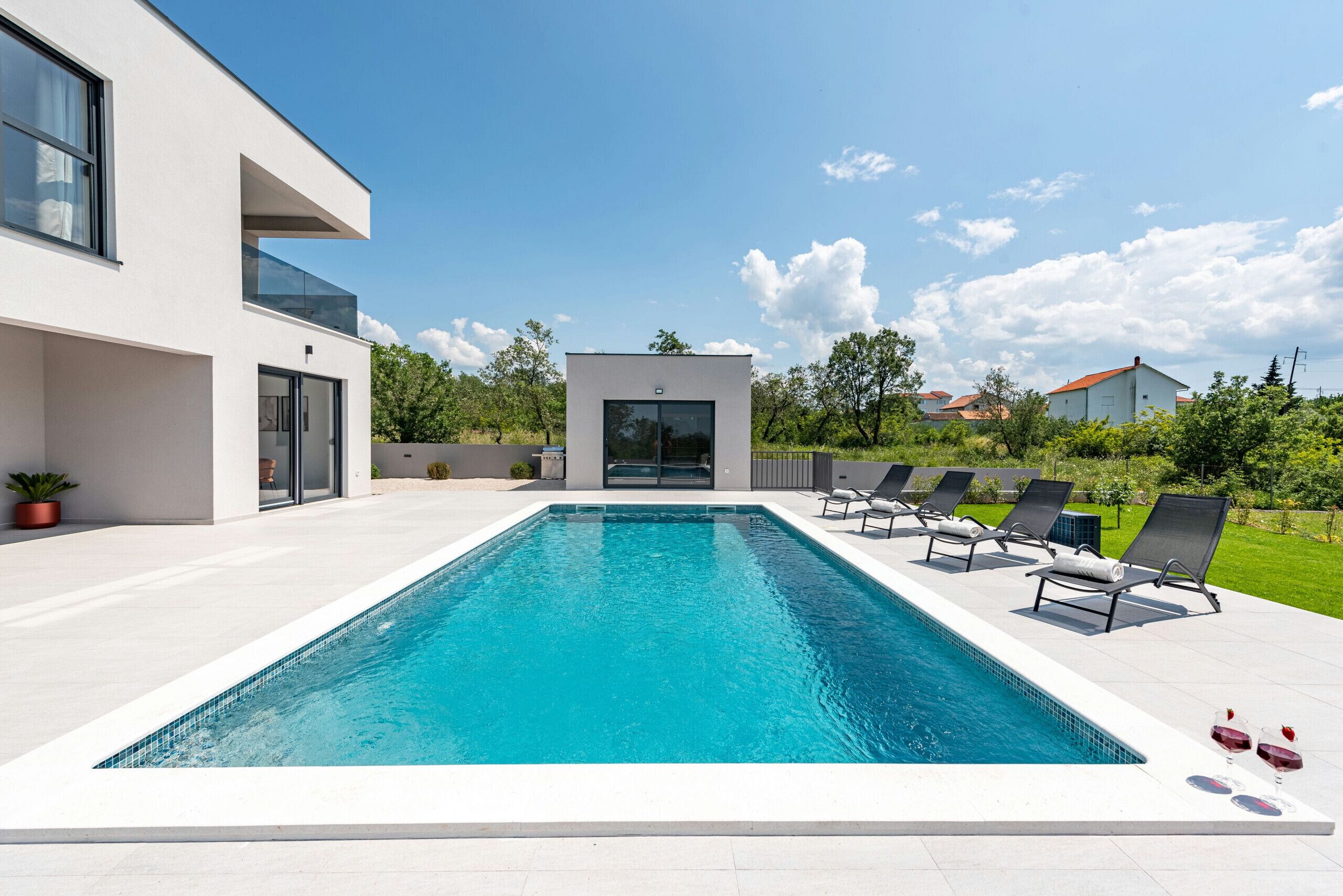 A modern pool area with a rectangular pool, lounge chairs, and a building with large windows and a glass door.