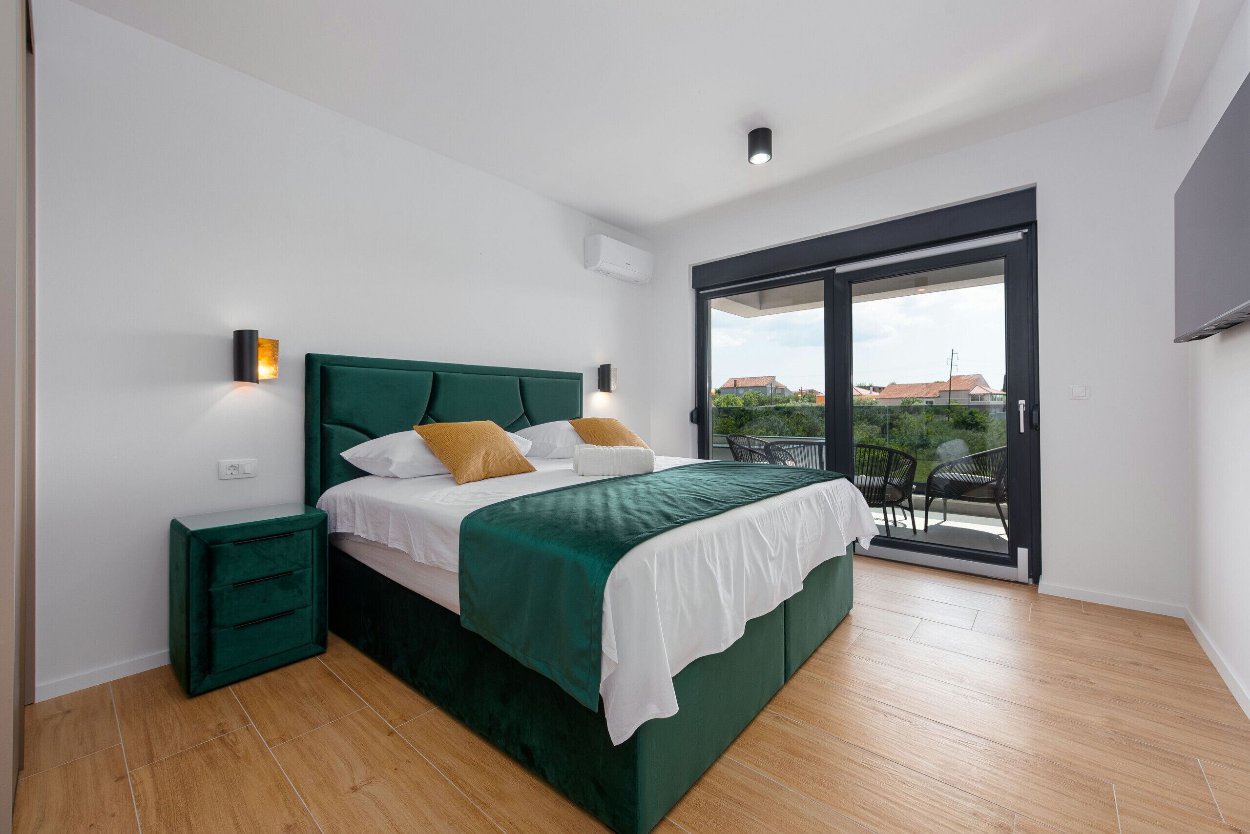 Camera da letto con un letto verde, biancheria bianca e vista sull'esterno attraverso una porta scorrevole in vetro.