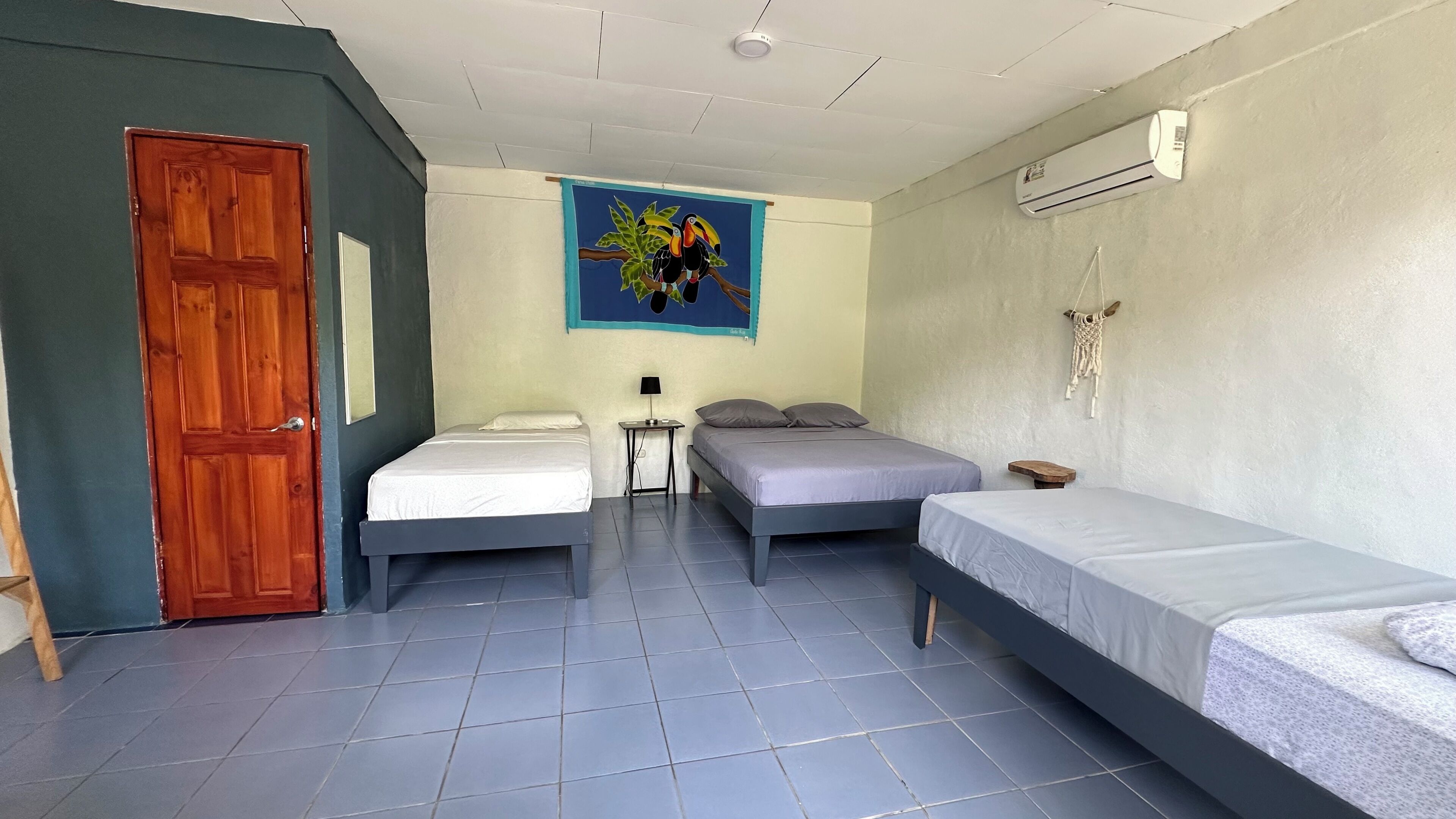 Quarto com duas camas de solteiro, uma mesa pequena com abajur, ar-condicionado fixado na parede e uma porta.