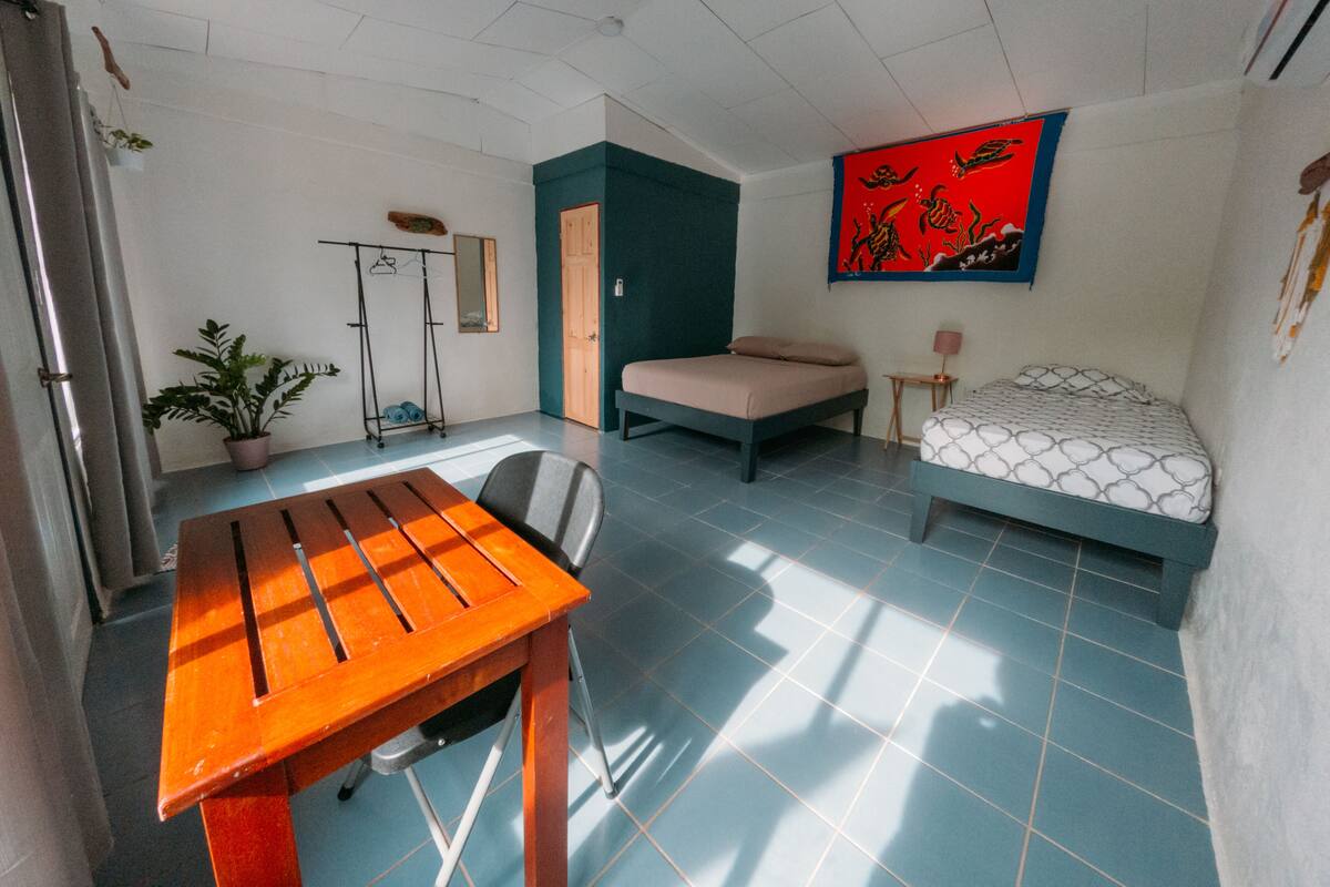 A compact room with a bed, a small table, a chair, a potted plant, and a wall with a colorful artwork.