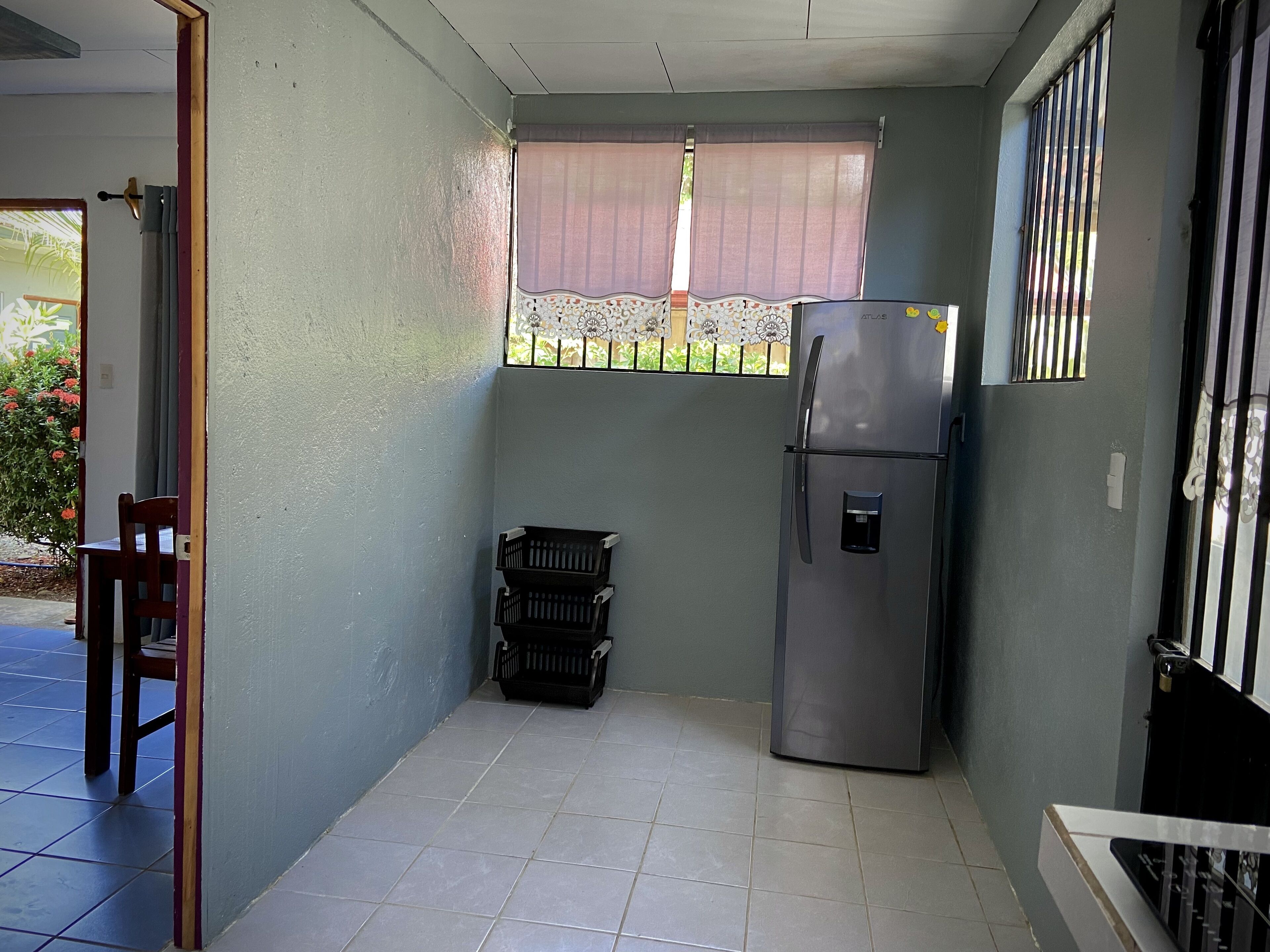 Cocina compacta con refrigerador, una ventana con celosía decorativa y una puerta que da a un espacio exterior.