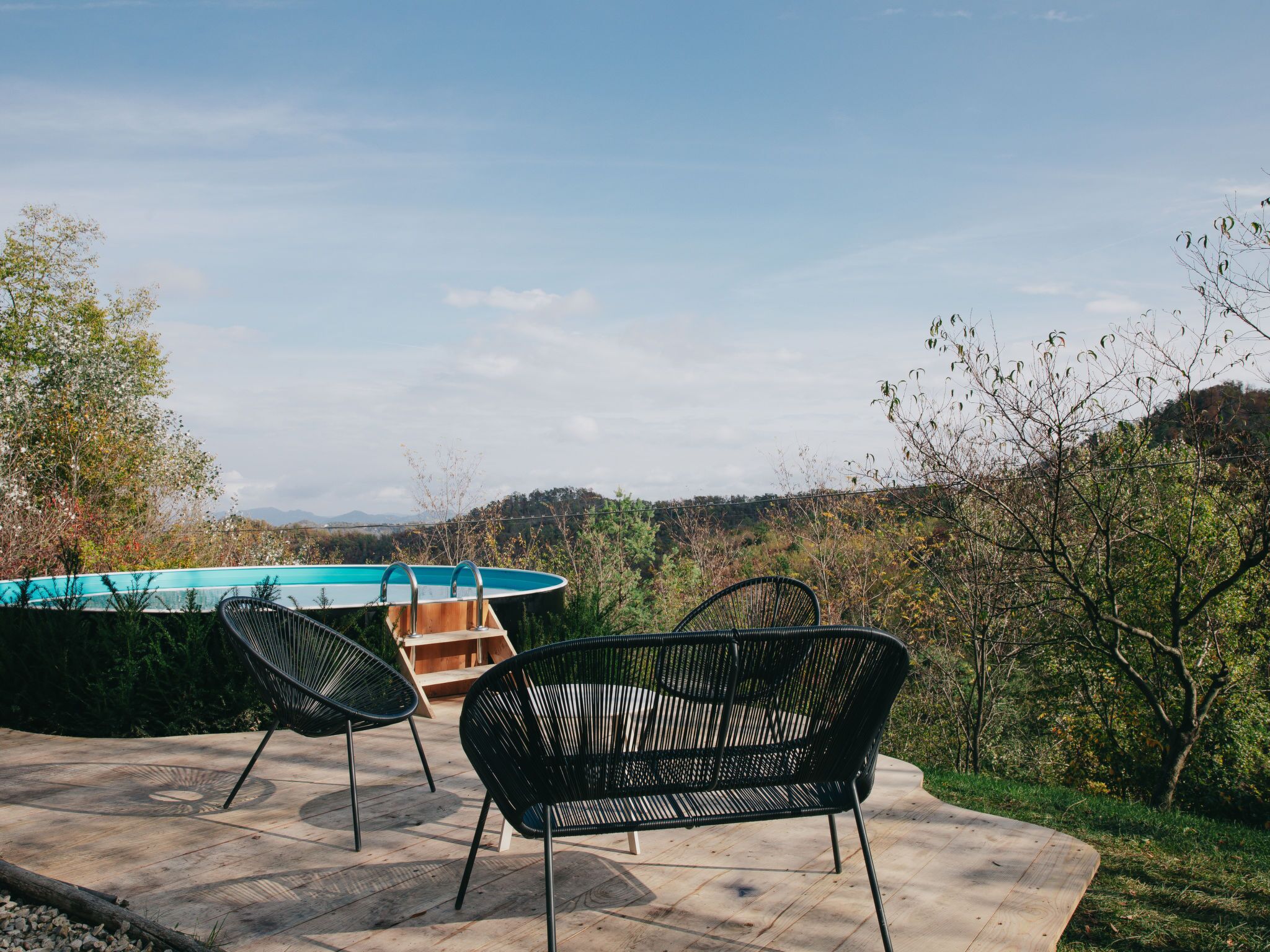 A pool area with two black wicker chairs and a wooden bench, surrounded by trees and a view of hills.