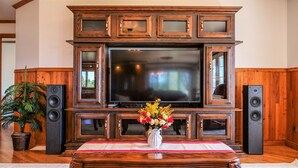 Una sala de estar con una gran televisión de pantalla plana montada en un mueble de madera, flanqueada por dos altavoces, una mesa de centro con un jarrón de flores y una planta en maceta.