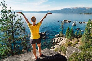 En person står på en klippa med armarna lyfta och blickar ut över en sjö, med berg i bakgrunden.