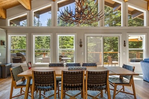 A spacious dining area with a large wooden table and chairs, large windows with a view of the outdoors, and a unique antler chandelier.
