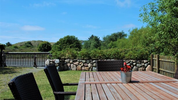 Une terrasse extérieure avec une table en bois et des chaises en osier, entourée d’un mur de pierre et d’une clôture en bois.