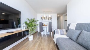 Una sala de estar moderna con un sofá gris, un comedor con una mesa y sillas blancas, y un ventanal grande.