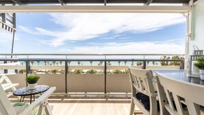Un balcón con vista a la playa, suelo de madera y muebles blancos.