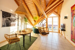 Relax in this stylish room with a round table, wicker chairs, and a hanging chair, featuring a large window and a beautiful painting.