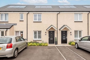 A two-story residential building with a beige exterior, black doors, and white window frames. There are two cars parked in the driveway, and the building has a flat roof with solar panels. - Fort Avenue (No 31) | Guardbridge (Leuchars)