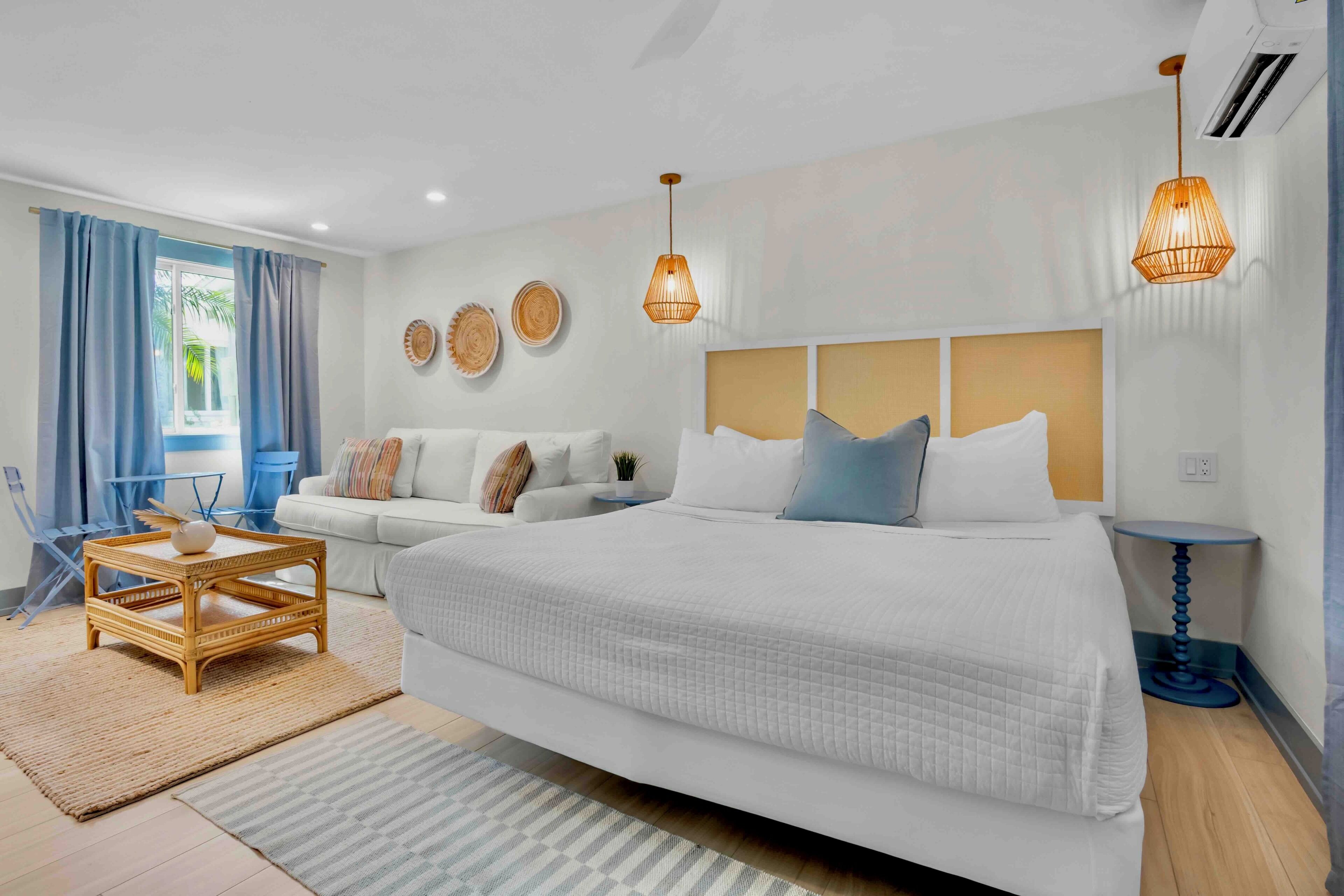 A modern bedroom with a large bed, a sofa, a small table, and a chair. There are wall decorations and two pendant lights.