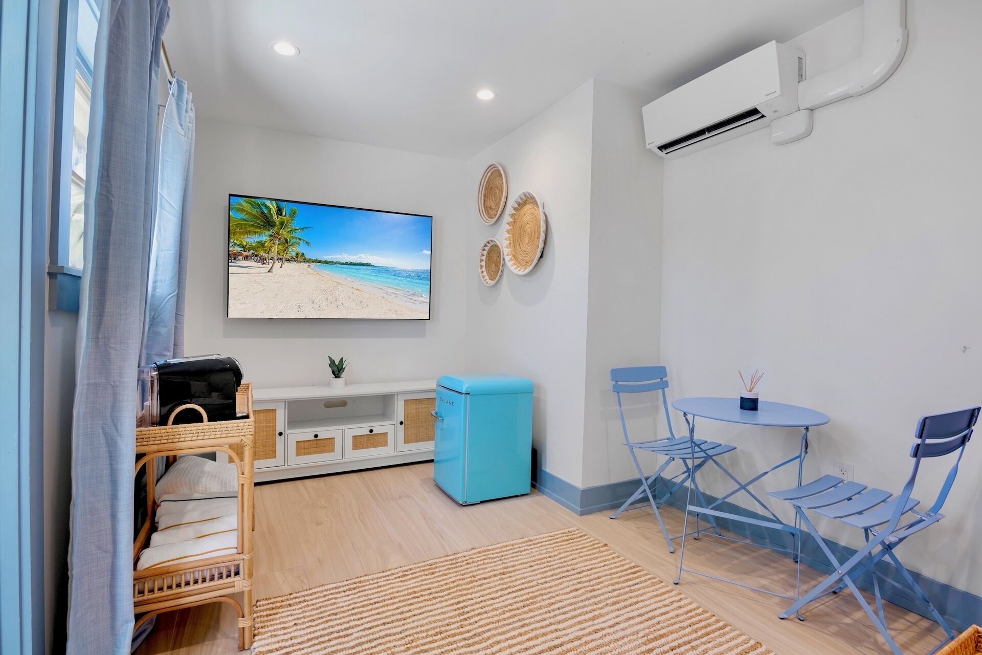 Ein Zimmer mit Fernseher, der eine Strandansicht zeigt, einem blauen Sofa, einem kleinen Tisch mit Stühlen und einer Klimaanlage.