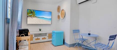A room with a beach scene TV, a blue sofa, a small table with chairs, and an air conditioning unit.