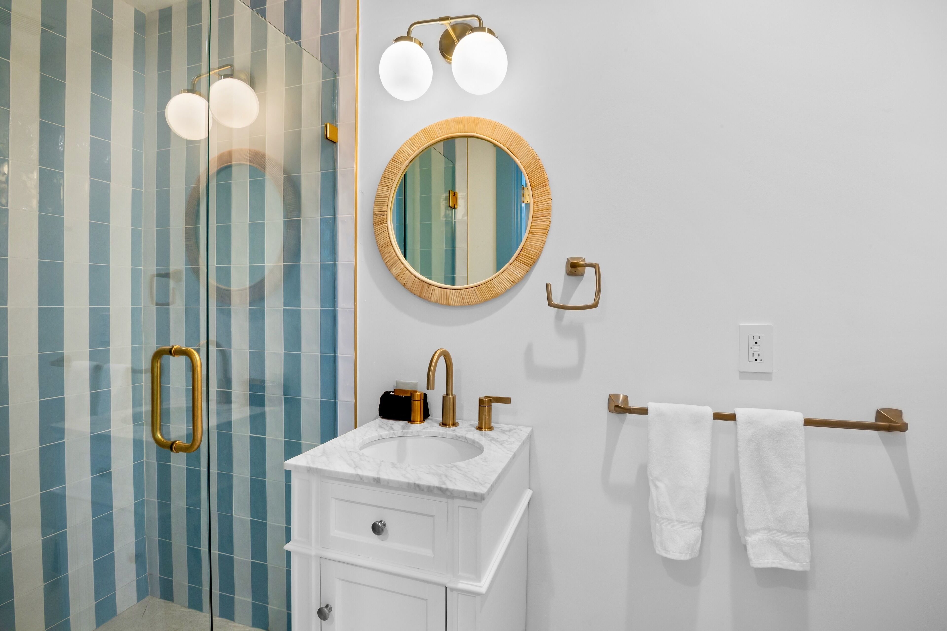 A bathroom with a glass shower enclosure, a white vanity with a marble sink, a round mirror with a wooden frame, and two towels hanging on a towel rack.
