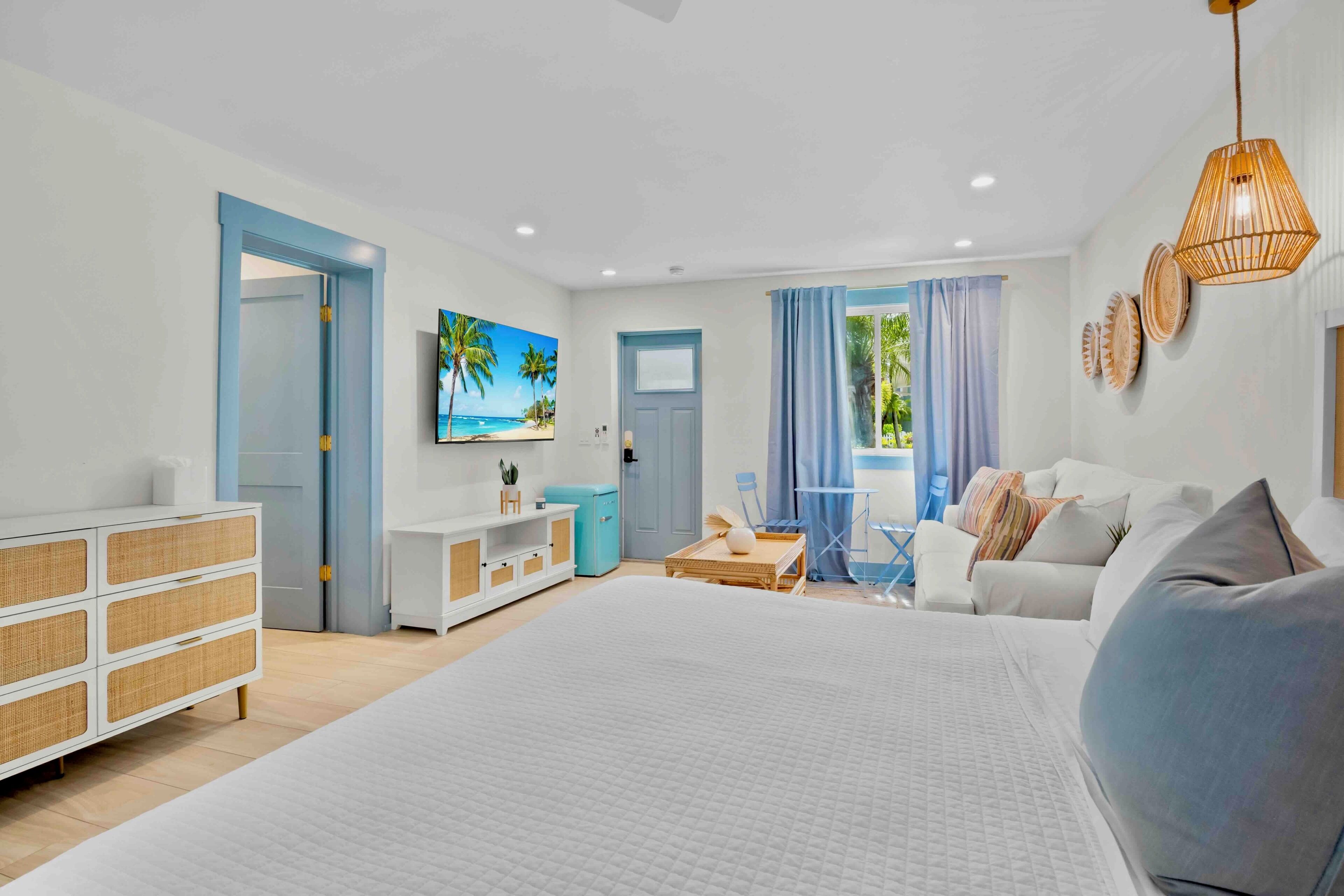 A modern bedroom with a large bed, a white dresser, a small table, and a blue door.