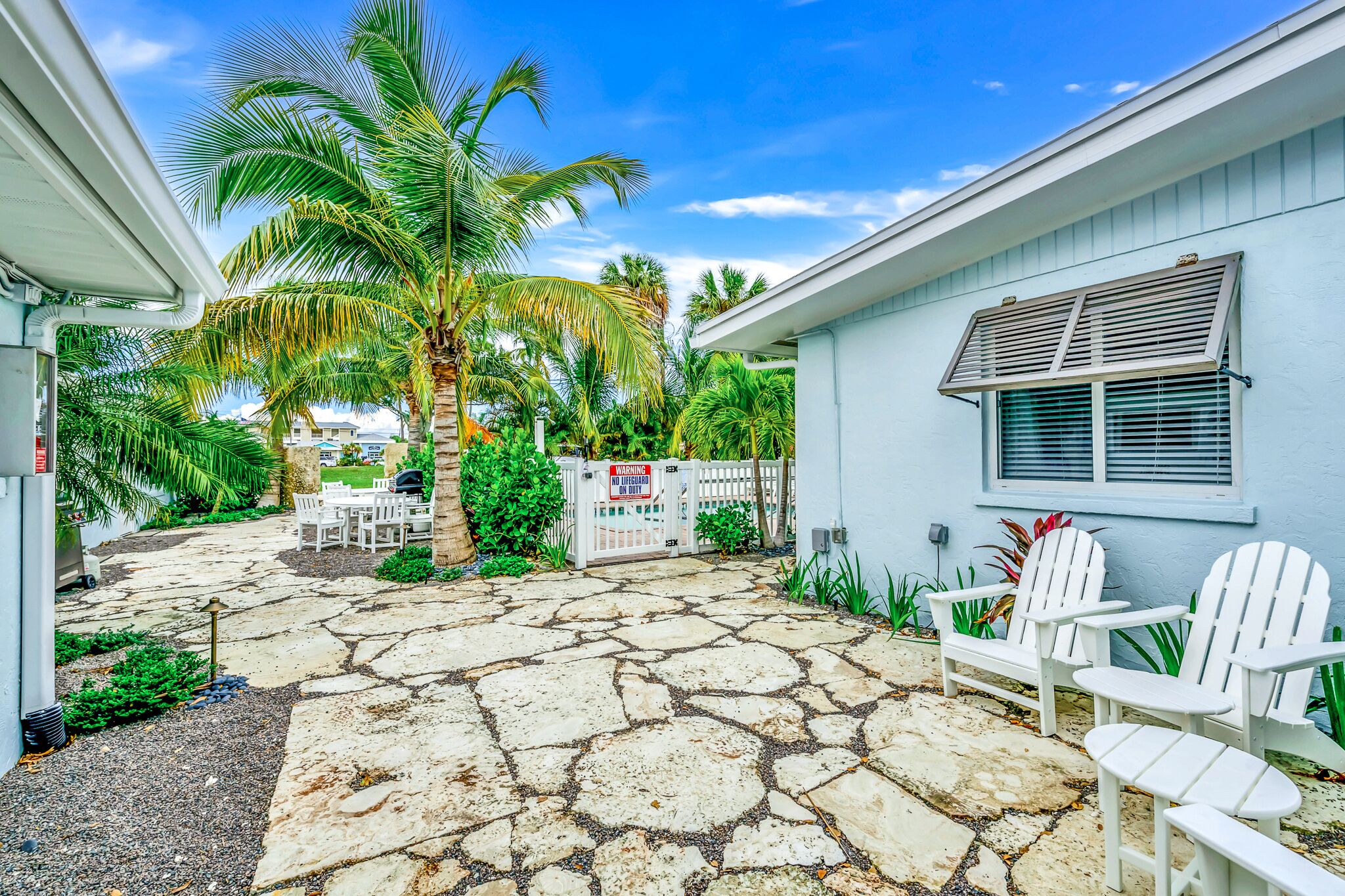 A house with a stone patio, white chairs, and a palm tree.