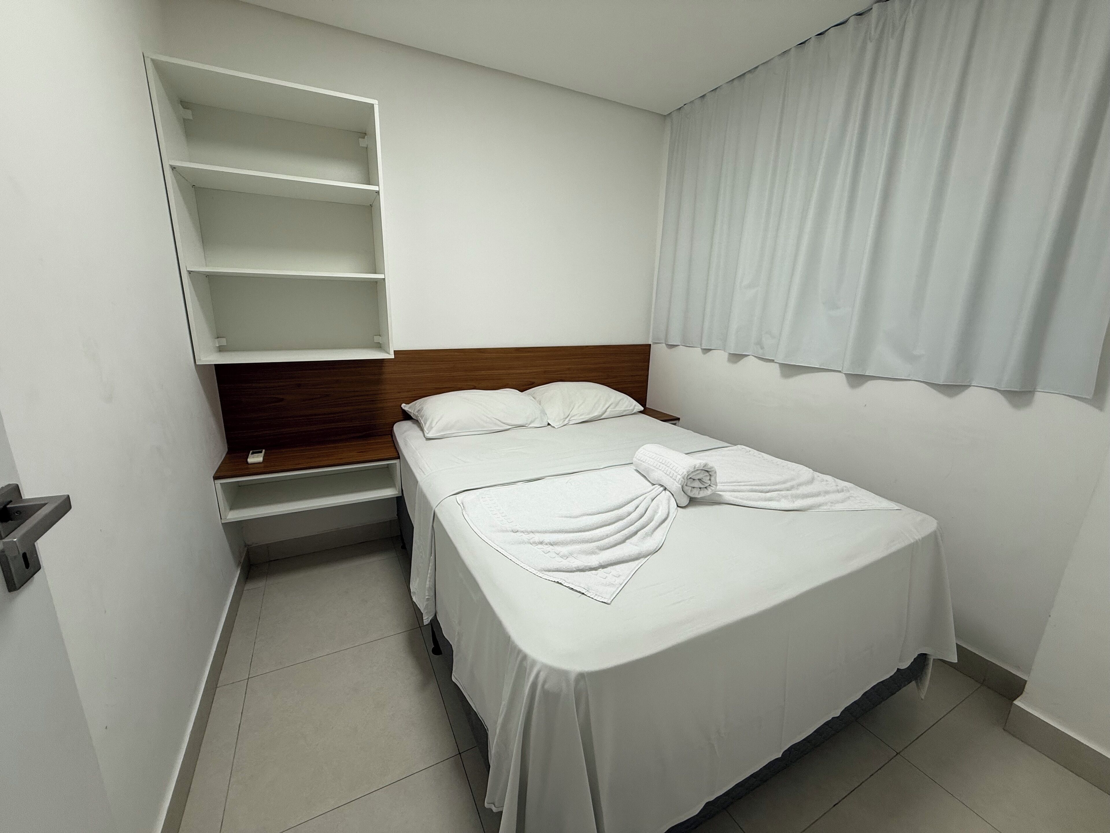 A single bed with white bedding, a wooden headboard, a built-in shelf unit, and a window with white curtains.