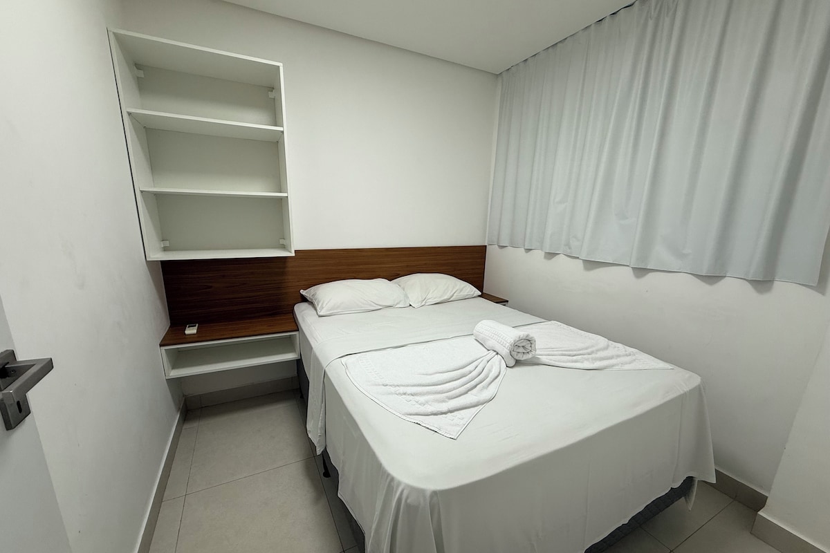 A single bed with white bedding, a wooden headboard, a built-in shelf unit, and a window with white curtains.