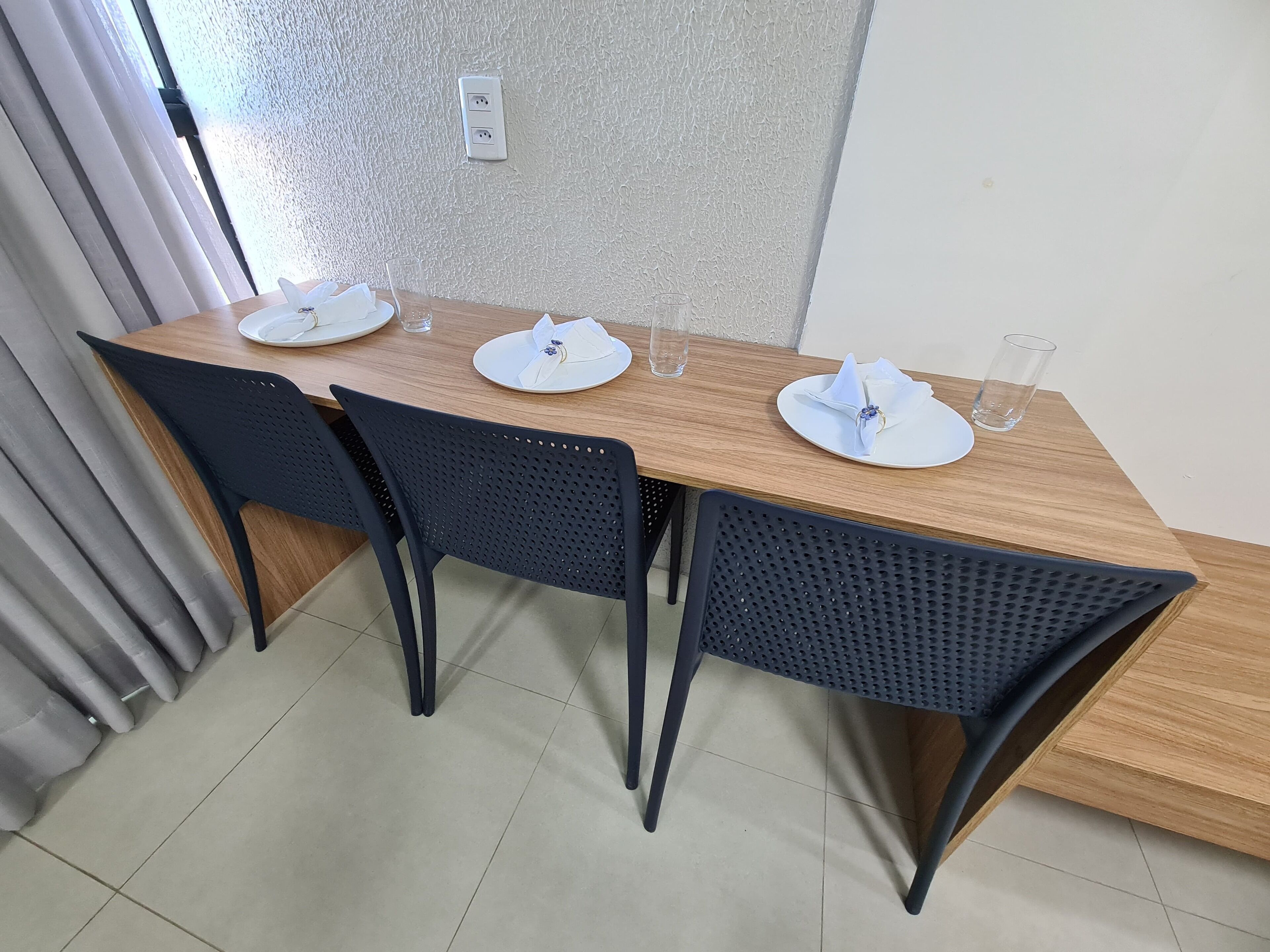 A dining table with four chairs, a power outlet on the wall, and a window with curtains.
