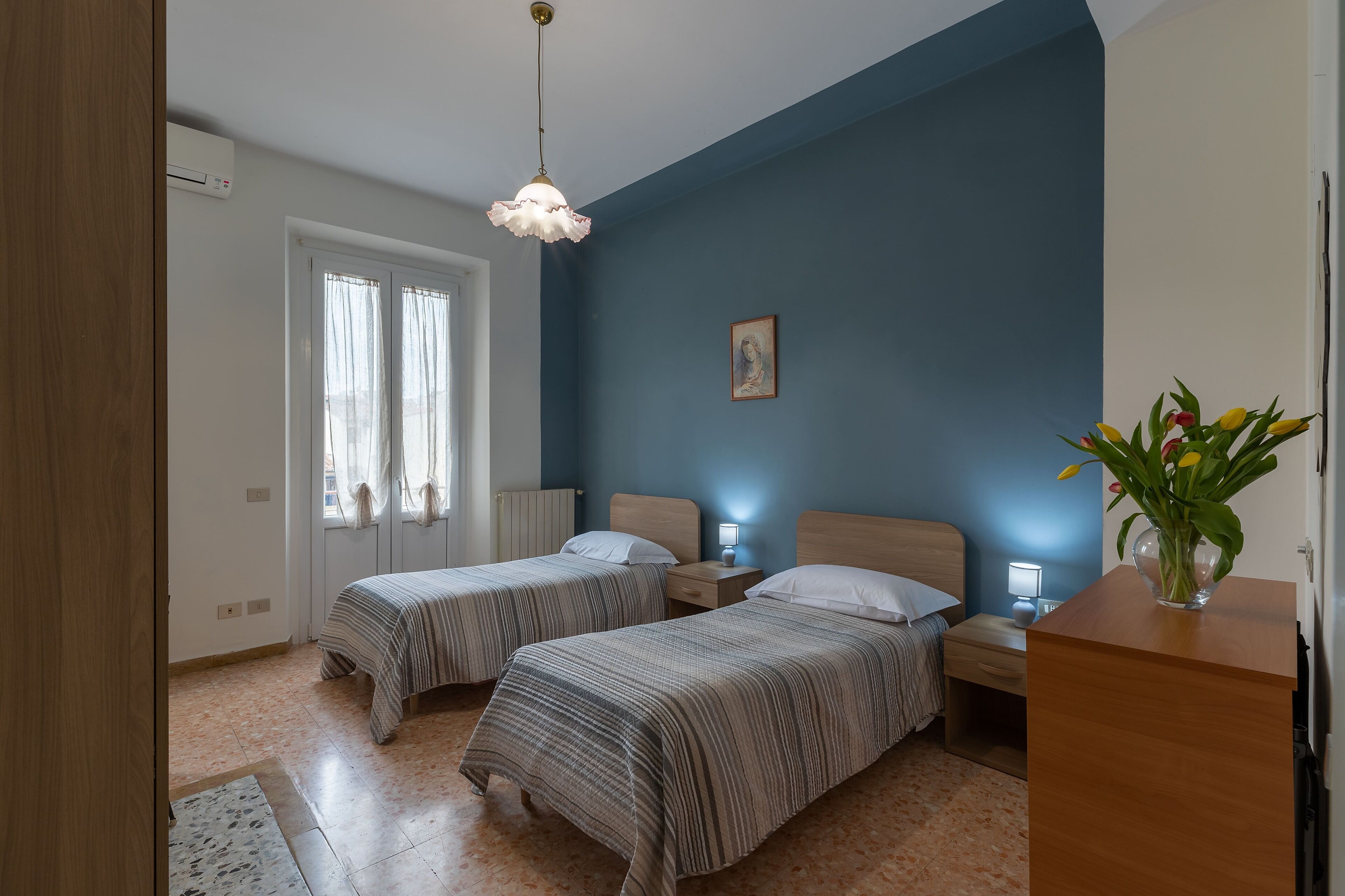 Un dormitorio con dos camas, una mesita de noche, un escritorio, un florero con flores y un cuadro en la pared.