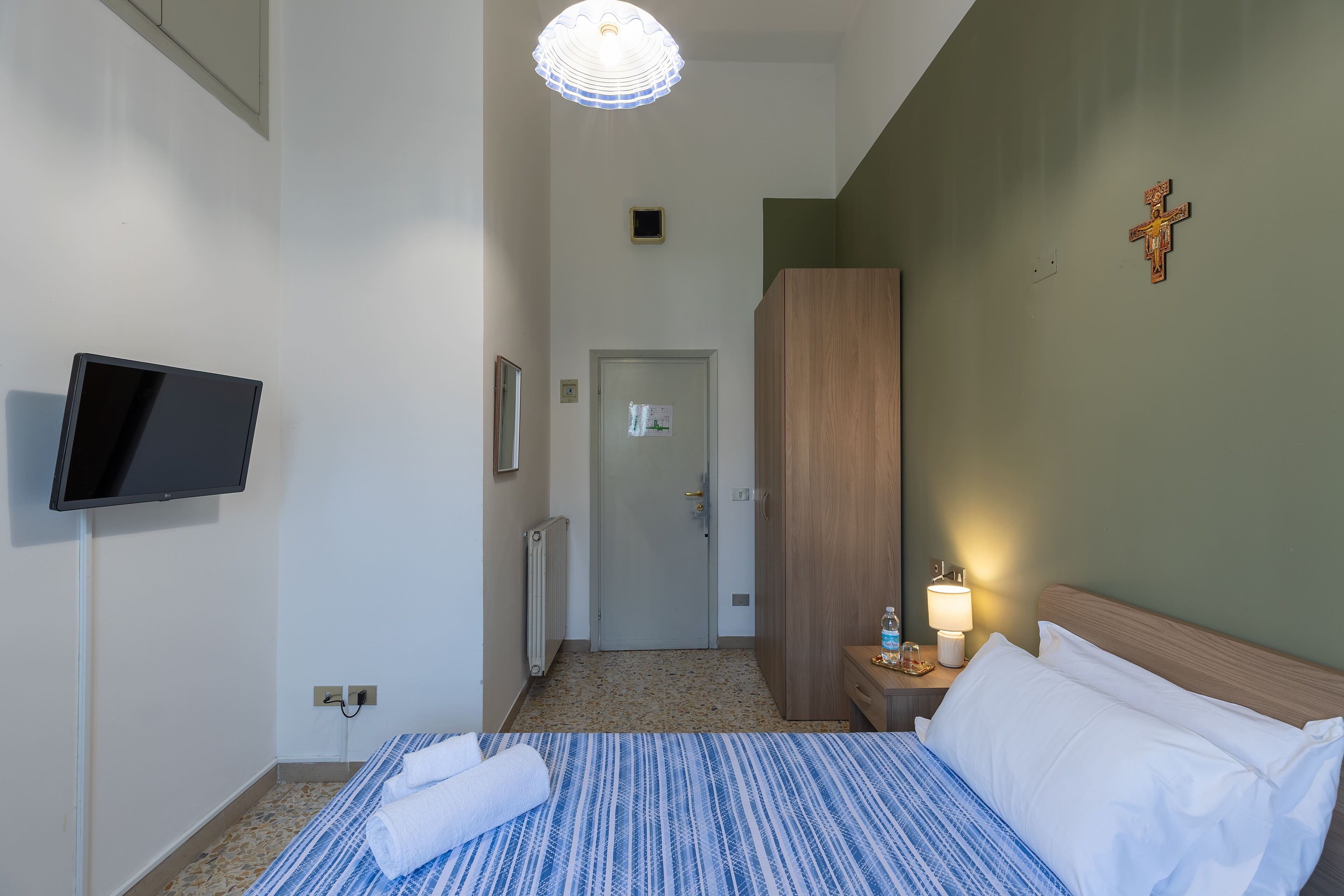 A hotel room with a bed, a television mounted on the wall, a wooden wardrobe, a bedside table with a lamp, and a cross on the wall.
