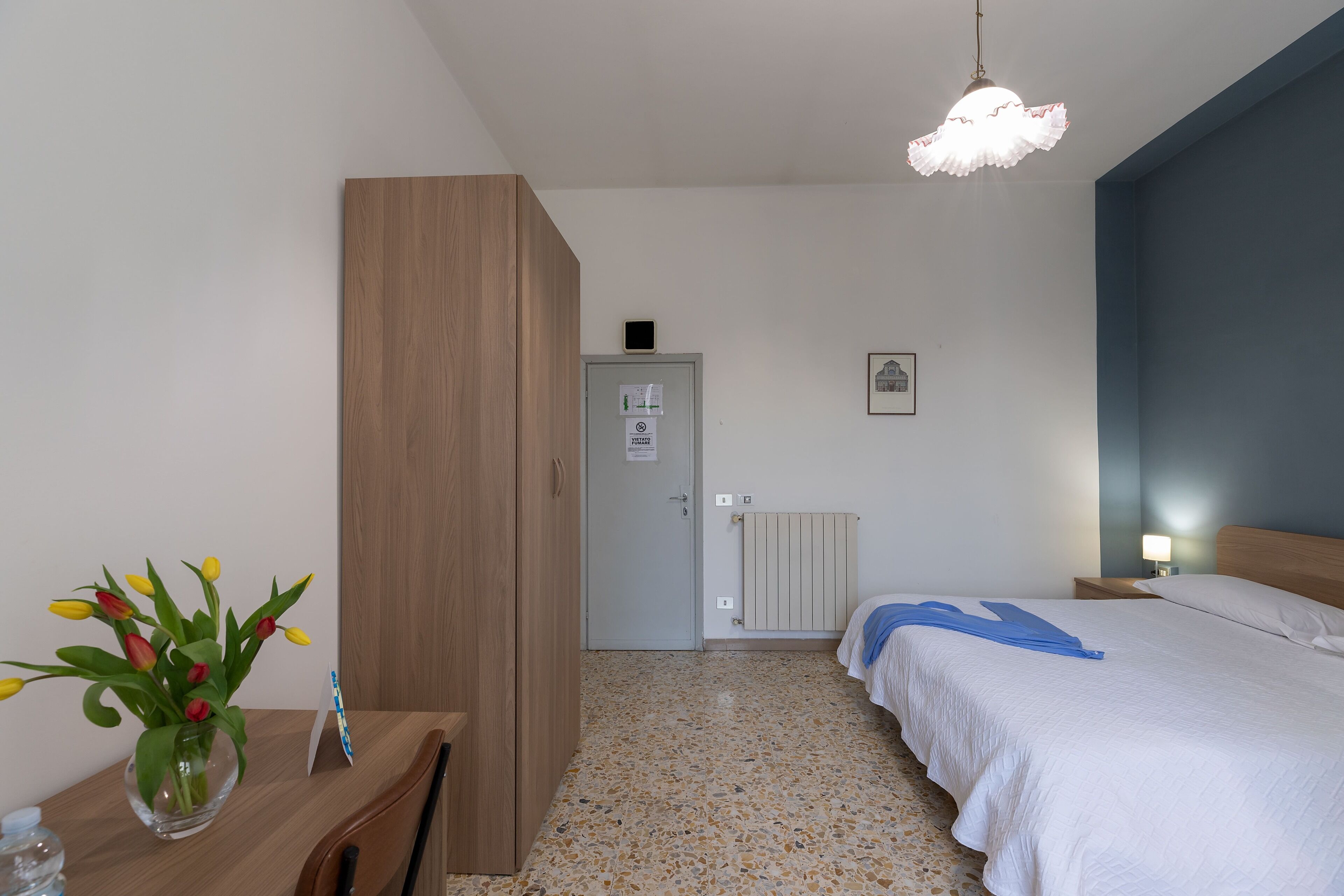 Une chambre d’hôtel avec un lit, un bureau agrémenté d’un vase de fleurs, une armoire, un radiateur et une porte avec une pancarte.