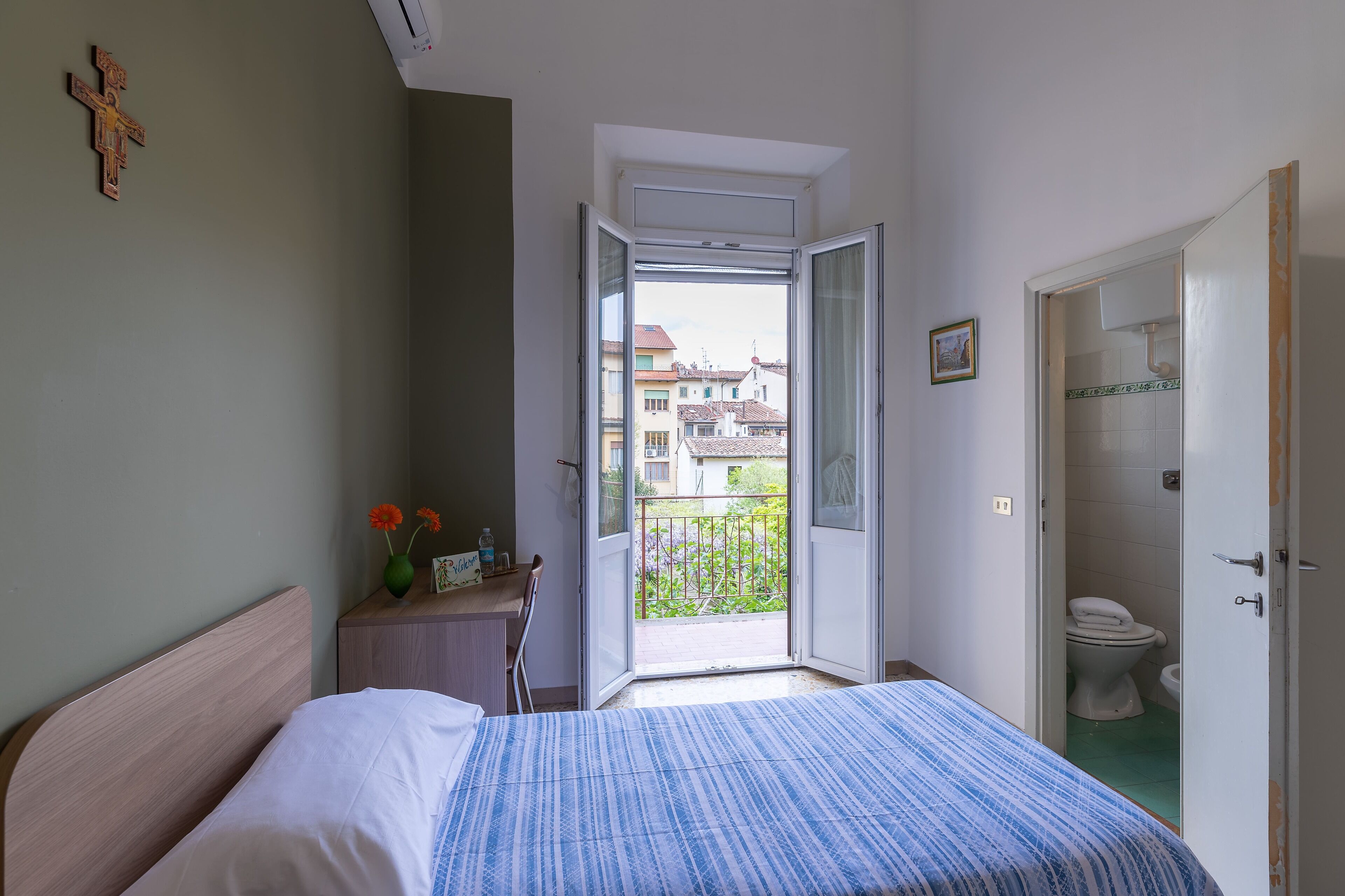 A bedroom with a bed, a small bedside table with a vase and a picture frame, a door leading to a balcony, and a view of buildings outside.