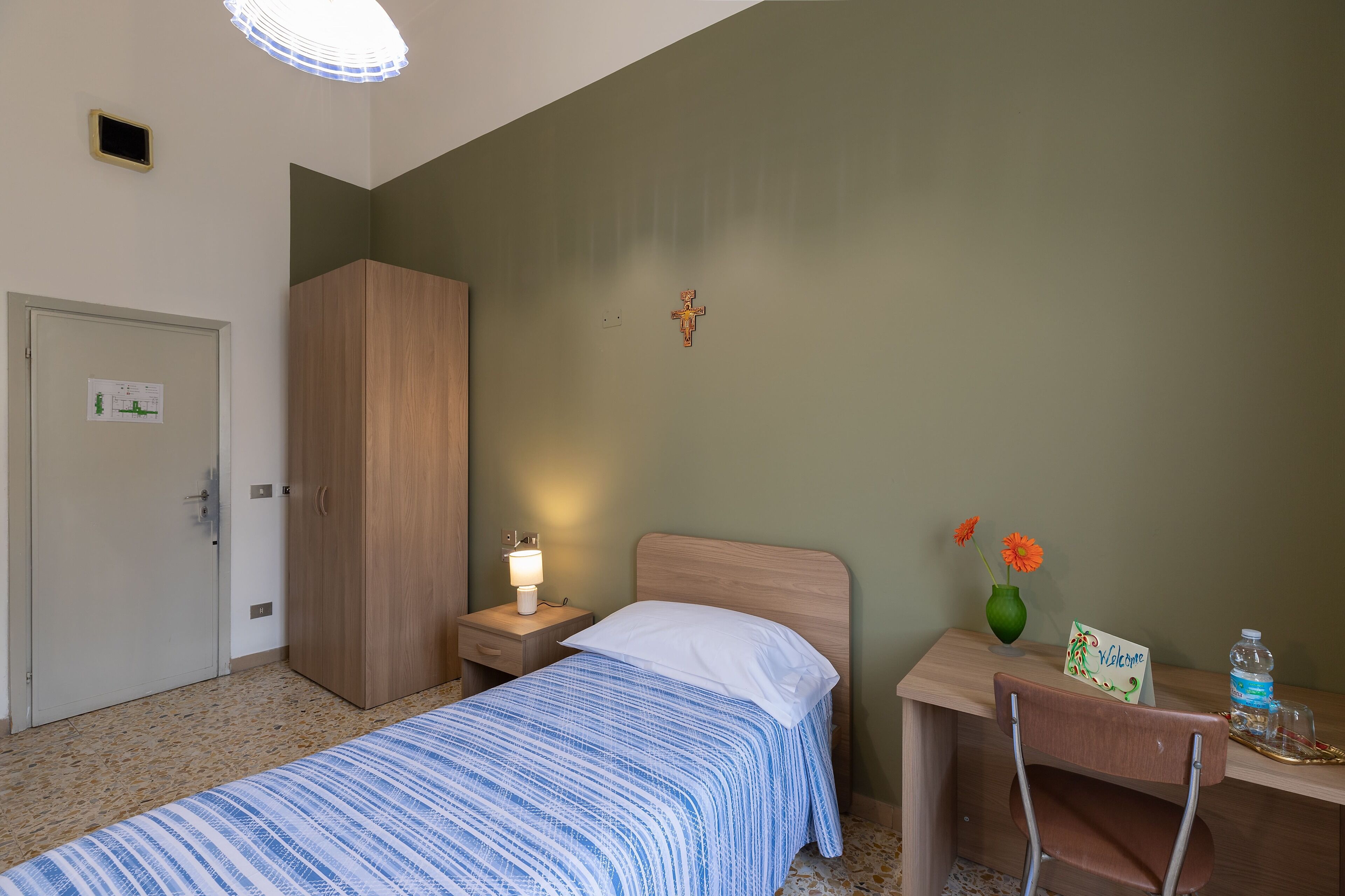 Une chambre à coucher avec un lit, une table de chevet, un bureau, une chaise, une armoire, une croix accrochée au mur et une pancarte de bienvenue.