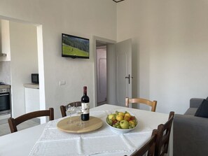Zona pranzo con tavolo apparecchiato per due, una ciotola di frutta, una bottiglia di vino e un televisore montato a parete.