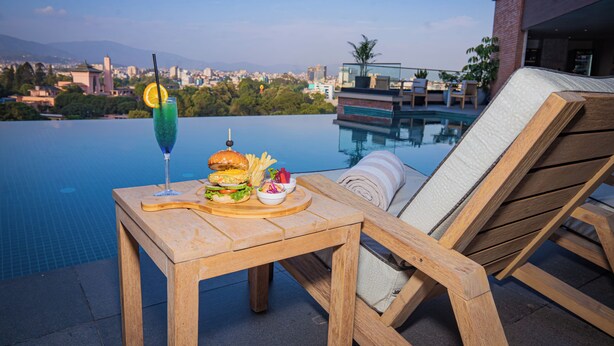 A wooden table with a burger and salad, a drink with a straw, and a chair by a pool with a city view in the background. - Hotel Barahi Kathmandu (Kathmandu)