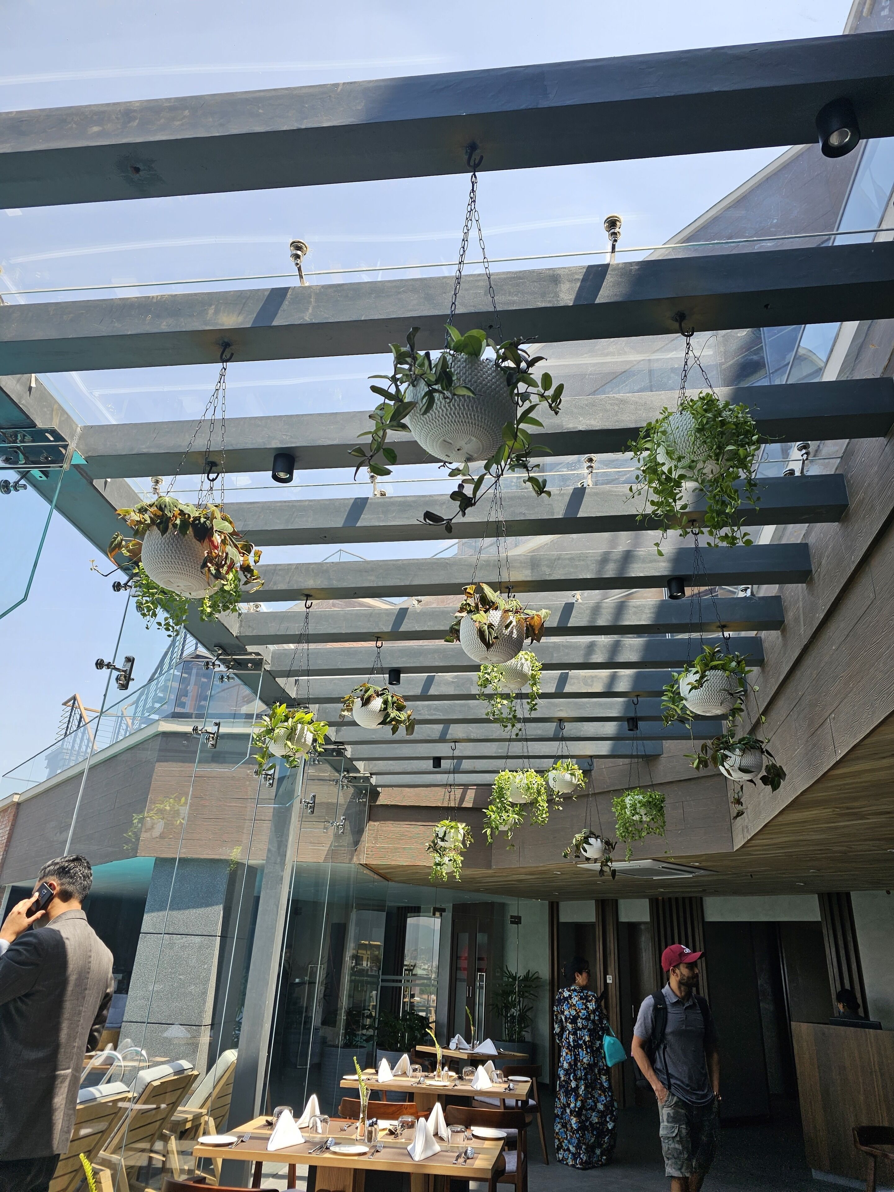 A modern outdoor dining area with multiple tables and chairs, surrounded by hanging planters with various plants.