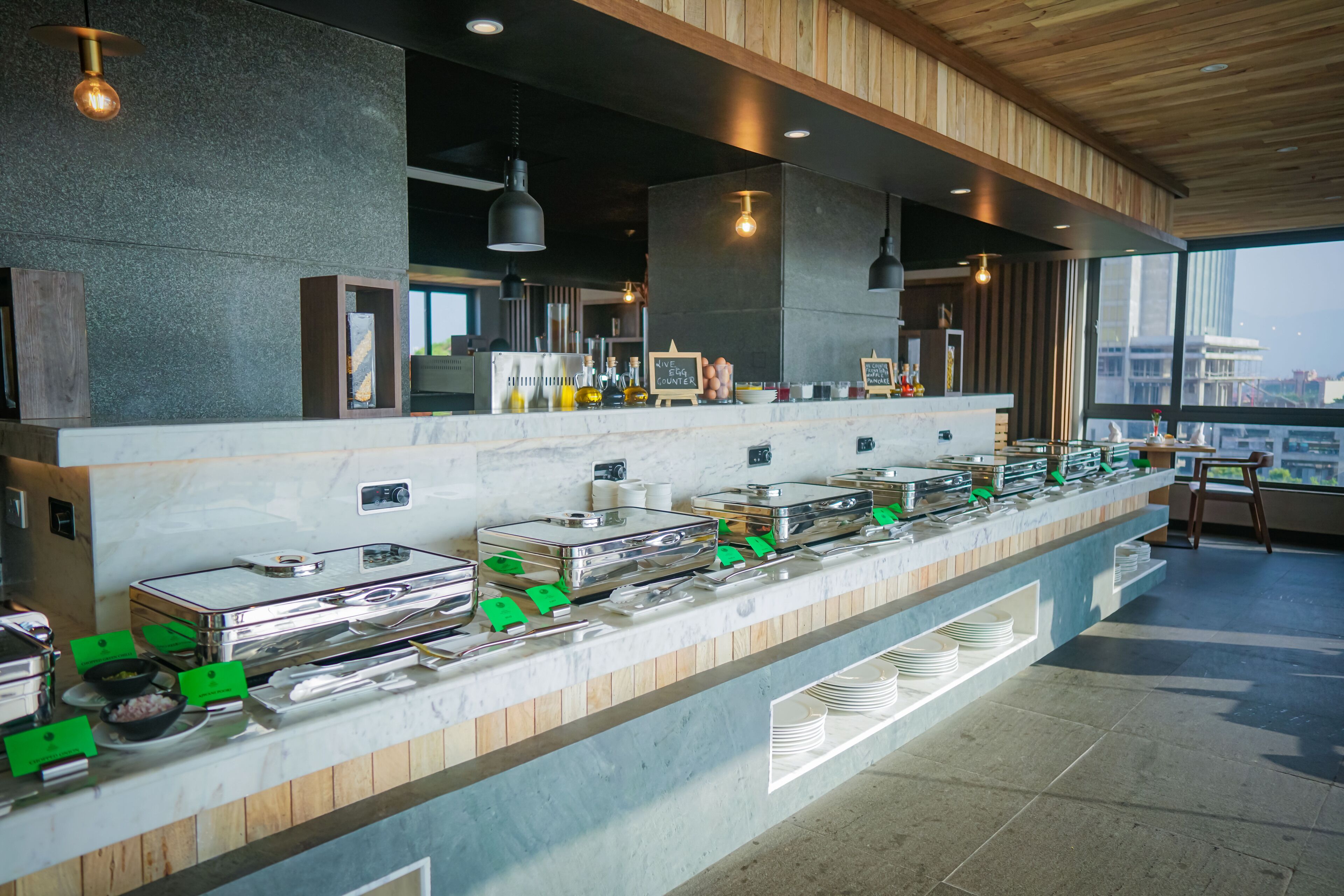 Disposição de buffet com diversas estações de comida, incluindo uma estação de pratos quentes e uma estação de frios, apresentando um design de interiores moderno e amplas janelas.